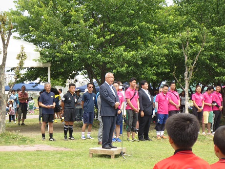 スカイパークの芝生の上で開会のあいさつをする三郷市長と、周りに立って話を聞く三郷JCカップ少年サッカー大会に出場する選手と関係者の写真