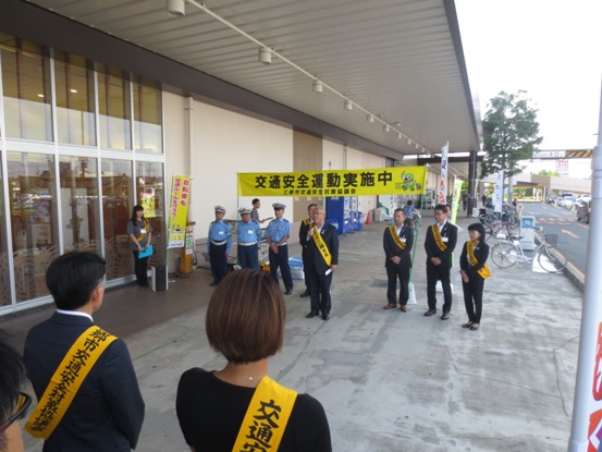 商業施設の建物外の通路に「交通安全運動実施中」と書かれた黄色地の横断幕が設置され、黄色のタスキを肩に掛けた方々や3名の警察官と4名の関係者が向かい合って立っている前で挨拶を行っている市長の写真