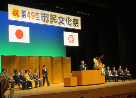 「祝 第49回市民文化祭」と書かれた横断幕と国旗と三郷市旗が飾られ金屏風が設置されたステージの演壇で挨拶を行っている市長の写真