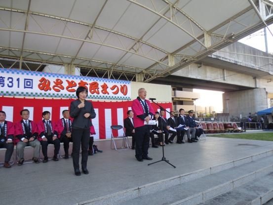 「第31回みさと市民まつり」と書かれた横断幕と紅白幕が飾られているにおどり公園のステージ上で、挨拶の言葉を述べるピンクの法被を着用した三郷市長と市長の横に立ち手話通訳をしている女性、後ろに置かれた席に座り話を聞く関係者の写真