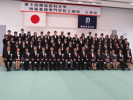 国旗と獨協医科大学の校旗の上に「第3回獨協医科大学附属看護専門学校三郷校入学式」と書かれた横断幕が掛けられ、赤と白の胸章を付けた三郷市長と関係者が入学生とともに記念撮影をしている写真
