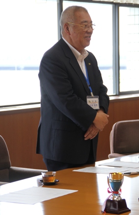 テーブルの上に小さなトロフィーが置かれ、市長が立って笑顔で挨拶を行っている写真
