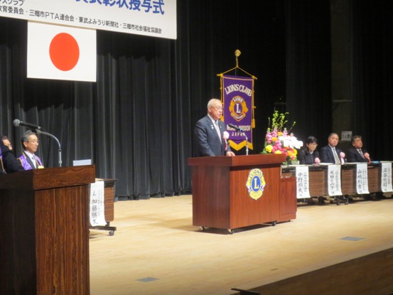 ライオンズクラブの旗が飾られている舞台上で、演壇に立って話をしている市長の写真