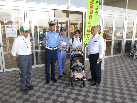 ピアラシティ玄関前で、三郷市長が関係者とともに「違法駐車・ポイ捨て」啓発のチラシを受け取ったベビーカーの子ども連れの女性に協力を呼び掛けている写真