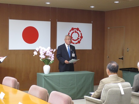 壁に国旗と三郷市旗が掲げられた壁を背に、薄ピンク色の花が入った花瓶が置かれた、緑色のクロスで覆われた演台に立った市長が、出席者に向かってあいさつの言葉を述べている写真