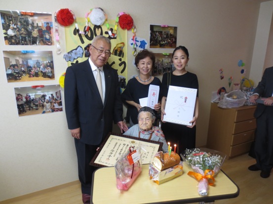 大倉ヨキさんのたくさんの写真と「100才おめでとう」と書かれた手作りのポスターが貼られている壁を背に、火の灯ったろうそくがさしてあるロールケーキや花束などが置かれたテーブルに座る表彰状を手に持った大倉ヨキさんと記念品を手に持ったご家族が三郷市長とともに写った記念写真
