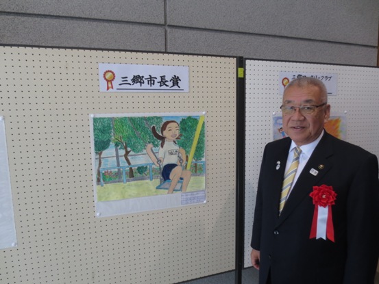 パネルに公園のブランコに乗って笑顔になっている女の子を描いた作品が展示され、作品の上には「三郷市長賞」の紙が貼られており、作品の横に並んで記念撮影をする市長の写真