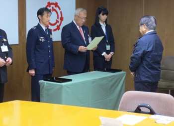 薄黄緑色のクロスを掛けたテーブルを挟んで男性と向かい合って立つ市長が委嘱状を読んでいる写真