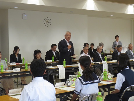 席に立ち資料を手に持ちながら話をする三郷市長と、席札とお茶のペットボトルが置かれた席に座り話を聞く関係者、市長や関係者と向かい合って座る各中学校の代表生徒たちの写真