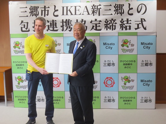黄色のTシャツを着たIKEAの関係者と市長が三郷市のバックボードの前で協定書を一緒に持って記念撮影をしている写真