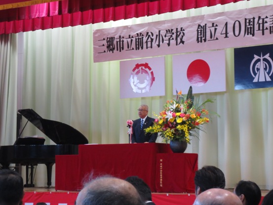 横断幕と国旗・三郷市旗・前谷小学校の校章旗が吊るしてあり、向かって左側にグランドピアノが置かれたステージの中央に置かれた、ピンクや黄色の豪華な花々で彩られた花台と並ぶ赤いカバーが掛けられた演台に立ち祝辞を述べる三郷市長の写真