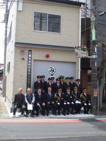 ベージュの2階建ての建物で、1階部分のシャッターの左側に「三郷市消防団第四分団第一班」と書かれた看板が設置されている機械器具置場の前で、三郷市長と関係者が並んで記念撮影をしている写真
