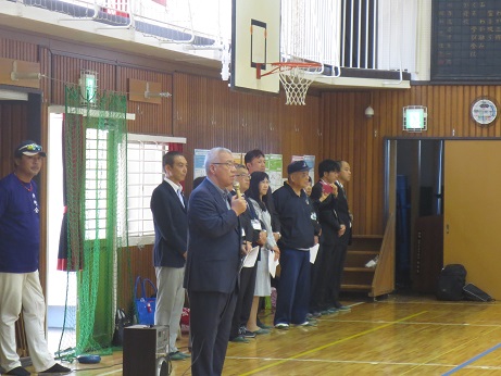 高州東小学校の体育館で、マイクを手に持ち挨拶の言葉を述べる三郷市長と バスケットゴールの下に横一列に並び話を聞く関係者の写真