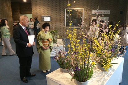 文化会館のロビーに黄色やピンク色、紫色などのカラフルな色で造られた生け花を、着物を着た女性と一緒に鑑賞している市長の写真