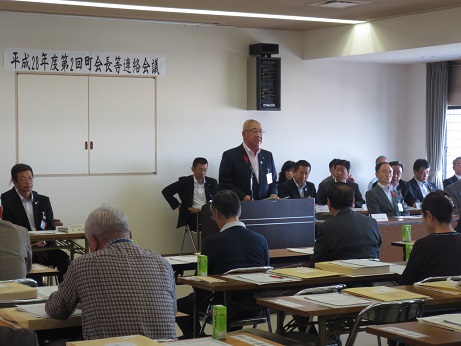 「平成28年度第2回町会長等連絡会議」と書かれた横幕が壁に貼られた室内で、左右の席に座っている関係者と参加者が向かい合って座り、中央の演壇に立ち挨拶を行っている市長の話を聞いている写真