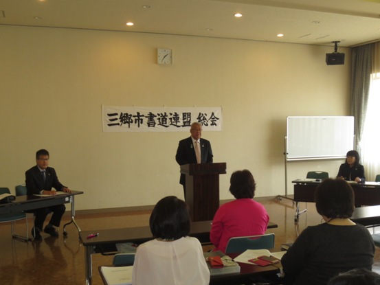 壁に「三郷市書道連盟総会」と書かれた紙が貼ってある部屋で、長テーブルに用意された椅子に着席している関係者と、演壇に立って挨拶をしている市長の写真