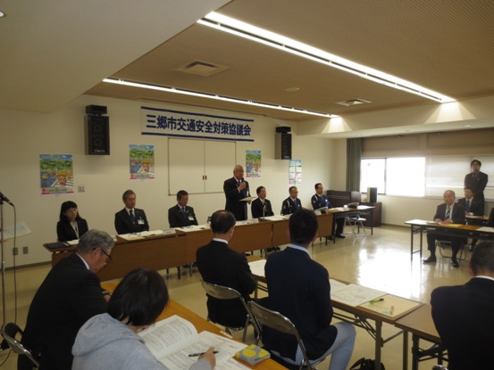壁に「三郷市交通安全対策協議会委員会」の横断幕が貼られた部屋で、長テーブルを囲んで多くの関係者が向かい合わせに座っている中、部屋の正面中央に立つ市長が話をしている写真