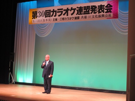「第30回カラオケ連盟発表会」と書かれた横断幕が吊るされているステージ上で、三郷市長が緑と青でライトアップされたスクリーンの前で挨拶の言葉を述べている写真