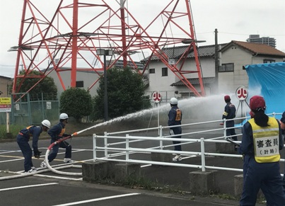 消防ホースを両手で持ち赤字で「火」と書かれたボードをめがけて放水している、紺の制服と白いヘルメットを着用しオレンジ色のゼッケンを付けた消防団員と、その様子を判定している黄色いゼッケンをつけた審査員の写真