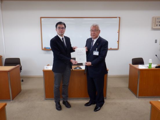 前方にホワイトボードが設置された会議室で、男性と三郷市長が委嘱書を一緒に持って記念撮影をしている写真
