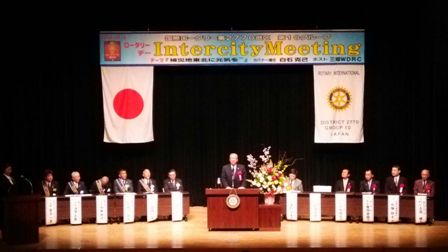 アルファベットでインターシティーミーティングと書かれた横断幕と国旗が飾られたステージの檀上で挨拶を行っている市長の写真