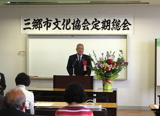 「三郷市文化協会定期総会」の紙が貼られた部屋で、等間隔に設置された長テーブルに参加者たちが着席しているなか、演壇に立っている市長が挨拶をしている様子の写真