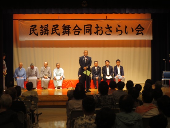 「民謡民舞合同おさらい会」の横断幕が設置されている舞台上で、赤いシートの敷かれた長椅子に座る関係者たちと、マイク台の前に立って挨拶をする市長の写真