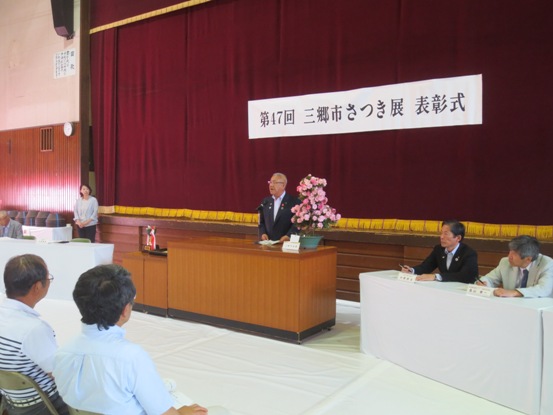 「第47回三郷市さつき展表彰式」のボードが設置された体育館で、ピンク色に咲くさつきの花の作品を横に置いた演壇で、市長が挨拶をしている写真
