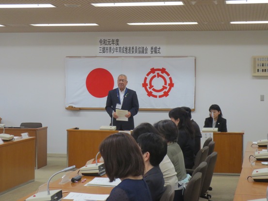 「令和元年度 三郷市青少年育成推進委員協議会委嘱式」の紙が壁に貼られている部屋で、椅子に着席している関係者たちと、正面中央で資料を手に挨拶をしている市長の写真
