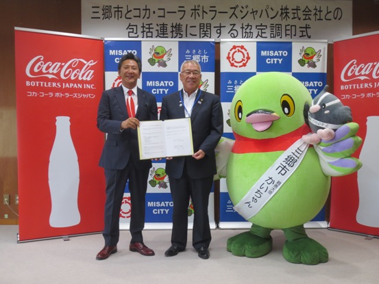 三郷市やコカ・コーラのバックボードの前で、市長と関係者が協定書を一緒に持っており、横に並ぶかいちゃんと一緒に記念撮影をしている写真