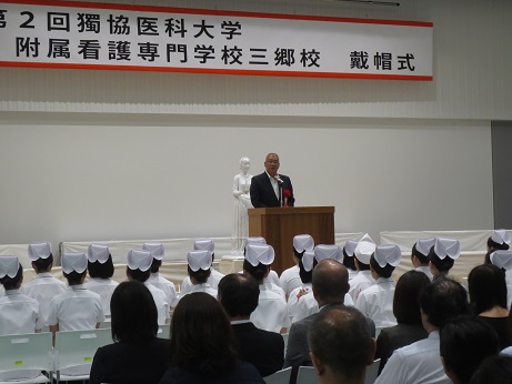 「第2階獨協医科大学附属看護専門学校三郷校戴帽式」と書かれた横断幕が壁に吊るしてあり、女性像の前に設置された演台に立ちあいさつの言葉を述べる三郷市長と、ナースウェアとナースキャップを着用し椅子に座って話を聞く学生の写真