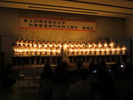 「第2階獨協医科大学附属看護専門学校三郷校戴帽式」と書かれた横断幕が吊るしてある壁の前で、ナースウェアとナースキャップを着用した学生たちが暗い会場の中、キャンドルを手に持ち並んでステージ台に立っている写真
