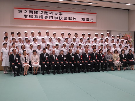 「第2階獨協医科大学附属看護専門学校三郷校戴帽式」と書かれた横断幕が吊るしてある壁の前で、胸章を付けた三郷市長と関係者がナースウェアとナースキャップを着用した学生たちとともに並び記念撮影をしている写真