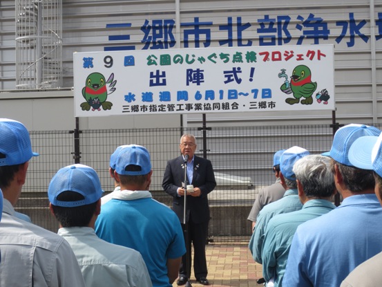 「第9回公園のじゃぐち点検プロジェクト出陣式」と書かれた横断幕の手前に設置されたマイクスタンドの前で挨拶を行っている市長の話を、向かい合って並んでいる水色の帽子を被った方々が聞いている写真