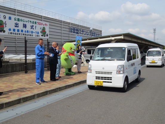 マスコットキャラクターかいちゃんや市長、関係者の方が並び拍手をするなか、軽ワゴン車に乗って公園の点検に出発する担当者を見送っている写真