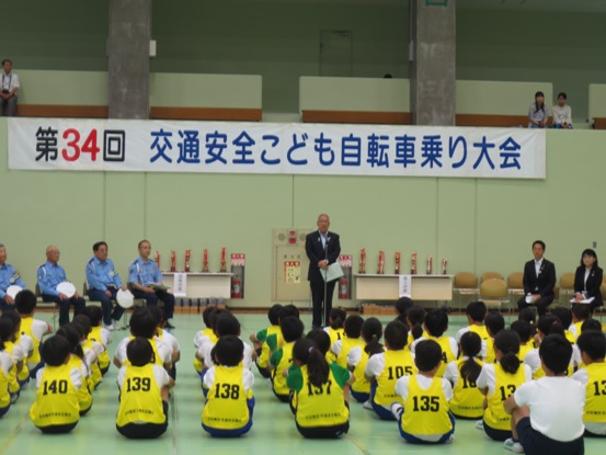 「第34回交通安全こども自転車乗り大会」と書かれた横断幕が壁に貼られ、壁際に設置された左右の長机の上に置いてあるトロフィーの前で挨拶を行っている市長の話を、黄色のゼッケンをつけた子供たちが床に体育座りをして聞いている後ろ姿の写真