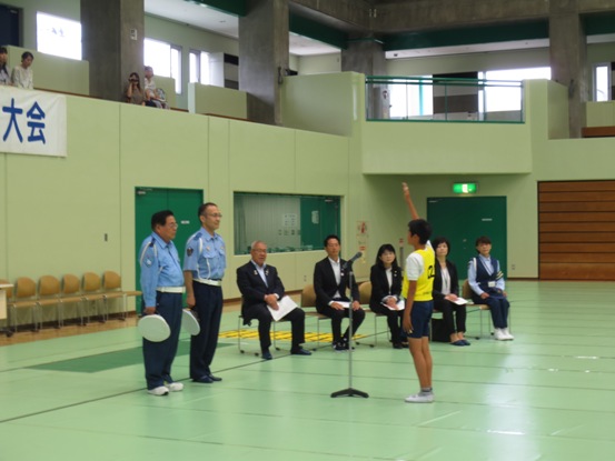 右奥に市長をはじめ5名の関係者が座り見守るなか、2名の警察官の方とマイクスタンドを挟んで黄色のゼッケンをつけた男の子が右手を真っ直ぐ上に上げ選手宣誓を行っている様子の写真