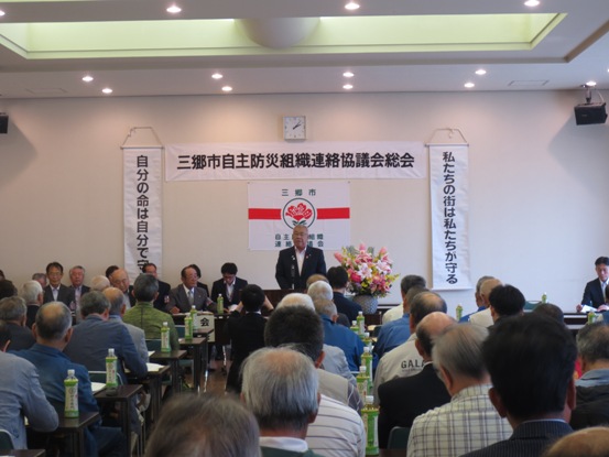 「三郷市自主防災組織連絡協議会総会」と書かれた横幕が貼られた室内で、関係者と参加者が向かい合って座り、中央の演壇で挨拶を行っている市長の写真