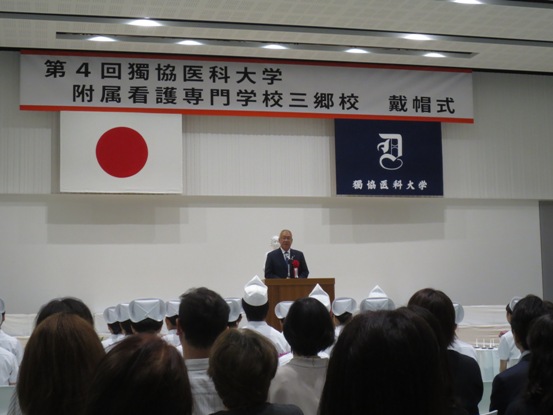 「第4回獨協医科大学附属看護専門学校三郷校戴帽式」と書かれた横断幕と国旗と校旗が飾られた下の演壇に立つ市長が、ナースキャップを被っている看護師の方々の前で挨拶を行っている様子の写真