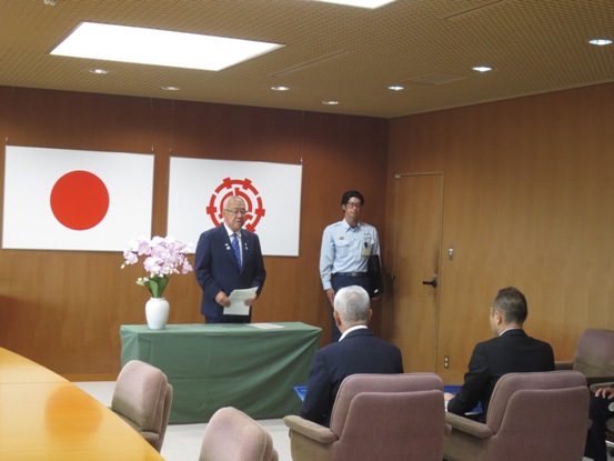 国旗と三郷市旗が飾られた室内で、椅子に座っている2名の会社の代表者と、薄黄緑色のクロスがかけられた台を挟んで向かい合って立っている市長が挨拶を行っている写真