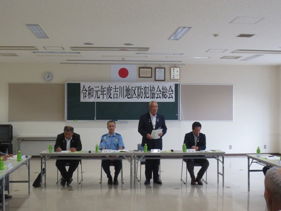 「令和元年度吉川地区防犯協会総会」と書かれた横幕が貼られた室内に設置された長机の席から立ち上がり挨拶を行っている市長の写真