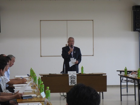 「会長」の役割名が貼られた席に立ち、あいさつの言葉を述べる三郷市長と、ペットボトルの置かれた席に座り資料に目を通しながら話を聞く参加者の写真