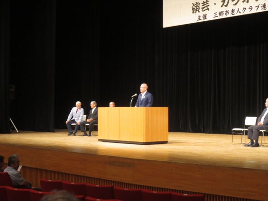 ステージ左右に設置された椅子に関係者が座り、中央の演壇で挨拶を行っている市長の写真