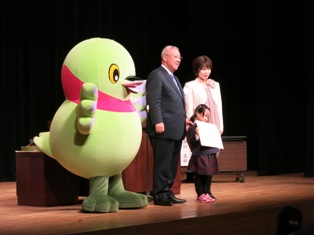 マスコットキャラクターかいちゃんと市長、女性と市長賞を受賞し賞状を持った女の子がステージの上に立っている写真