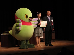 マスコットキャラクターかいちゃんと市長、市長賞を受賞し賞状を両手で持った膝丈のスカートを着た女性がステージの上に立っている写真