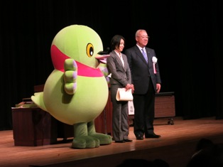 マスコットキャラクターかいちゃんと市長、市長賞を受賞し賞状を半分に曲げて持ったグレー色のスーツを着た女性がステージの上に立っている写真