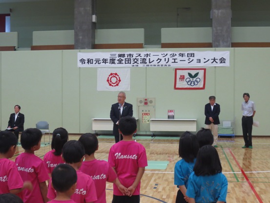 「三郷市スポーツ少年団令和元年度全団交流レクリエーション大会」と書かれた横断幕が壁に貼られ、体育館に設置されたマイクスタンドの前で挨拶を行っている市長の話を、ピンク色や水色のシャツを着た子供たちが並んで聞いている様子を後ろから写した写真