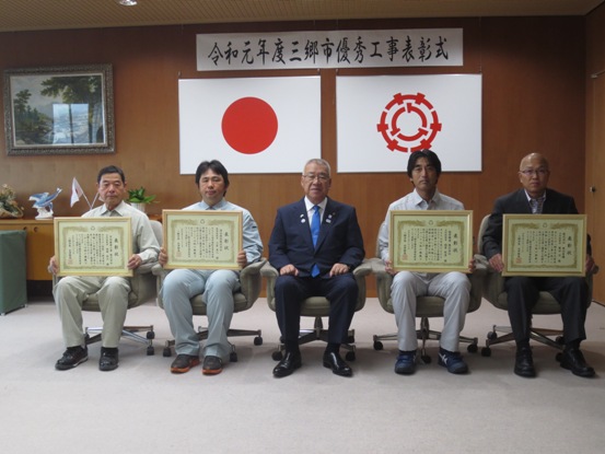 「令和元年度三郷市優秀工事表彰式」と書かれた横幕と国旗と三郷市旗が飾られた室内で、市長を中央にし、額入りの表彰状を持っている4名の受賞者が横一列に並んで椅子に座っている写真