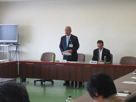 席に立ち資料を手に持ちながら挨拶の言葉を述べる三郷市長と、席に座り資料に目を通しながら話を聞く関係者の写真