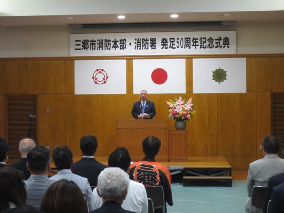「三郷市消防本部・消防署発足50周年記念式典」と書かれた横断幕と国旗と三郷市旗が飾られた会場に設置された椅子に座っている参加者と向かい合っている市長が、檀上で挨拶を行っている様子の写真